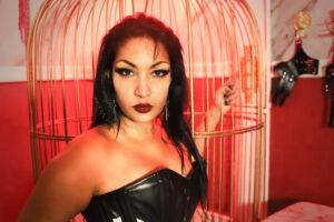October 2026 Dominatrix Scarlet Vexus next to a cage in her dungeon